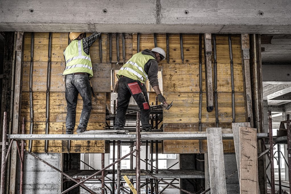 Zwei Bauarbeiter in Warnwesten stehen auf einem Gerüst und arbeiten an einer Betondecke