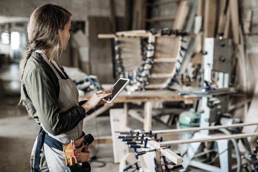 Eine Handwerkerin steht in einer Holzwerkstatt und hat ein Tablet in der Hand