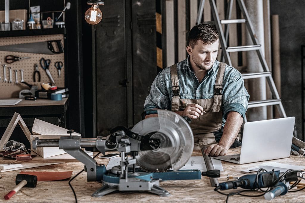 Ein junger Tischler sitzt in einer Holzwerkstatt und plant am Laptop sein nächstes Projekt